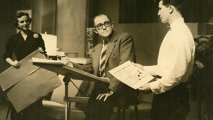 Three people in a vintage office or classroom setting: a woman standing on the left, a man seated at a desk with papers, and a young man standing on the right holding a comic book, all engaged in conversation or presentation.