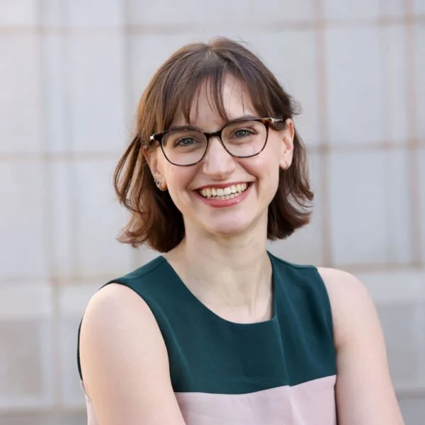 A woman with glasses and shoulder-length brown hair smiling at the camera, wearing a sleeveless top, standing outdoors with a light-colored tiled background.