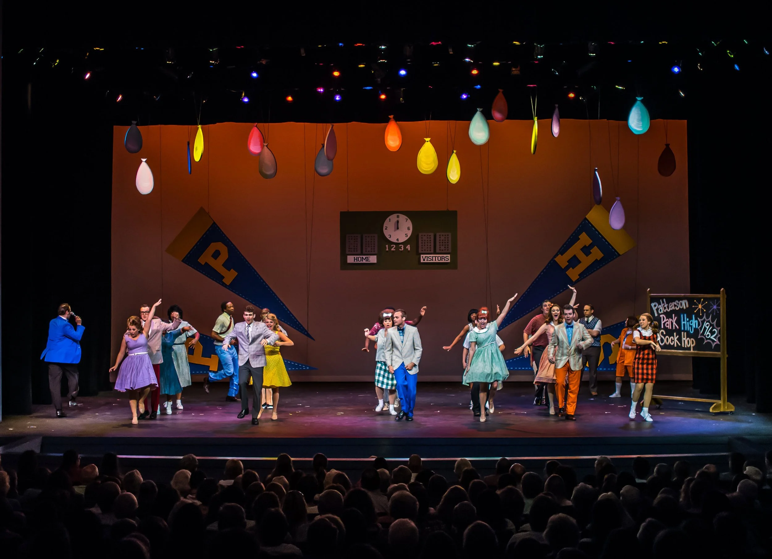 Stage performance of students at Patterson Park High School Sock Hop, with colorful balloons hanging from the ceiling and a scoreboard in the background.