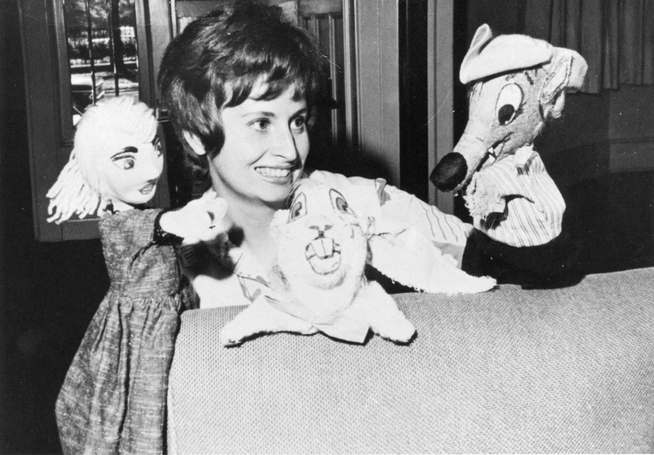 A woman smiling and sitting at a table with two puppets, one on her left with a doll face and the other on her right with a dog face, both wearing striped clothing. The background shows wooden cabinets.