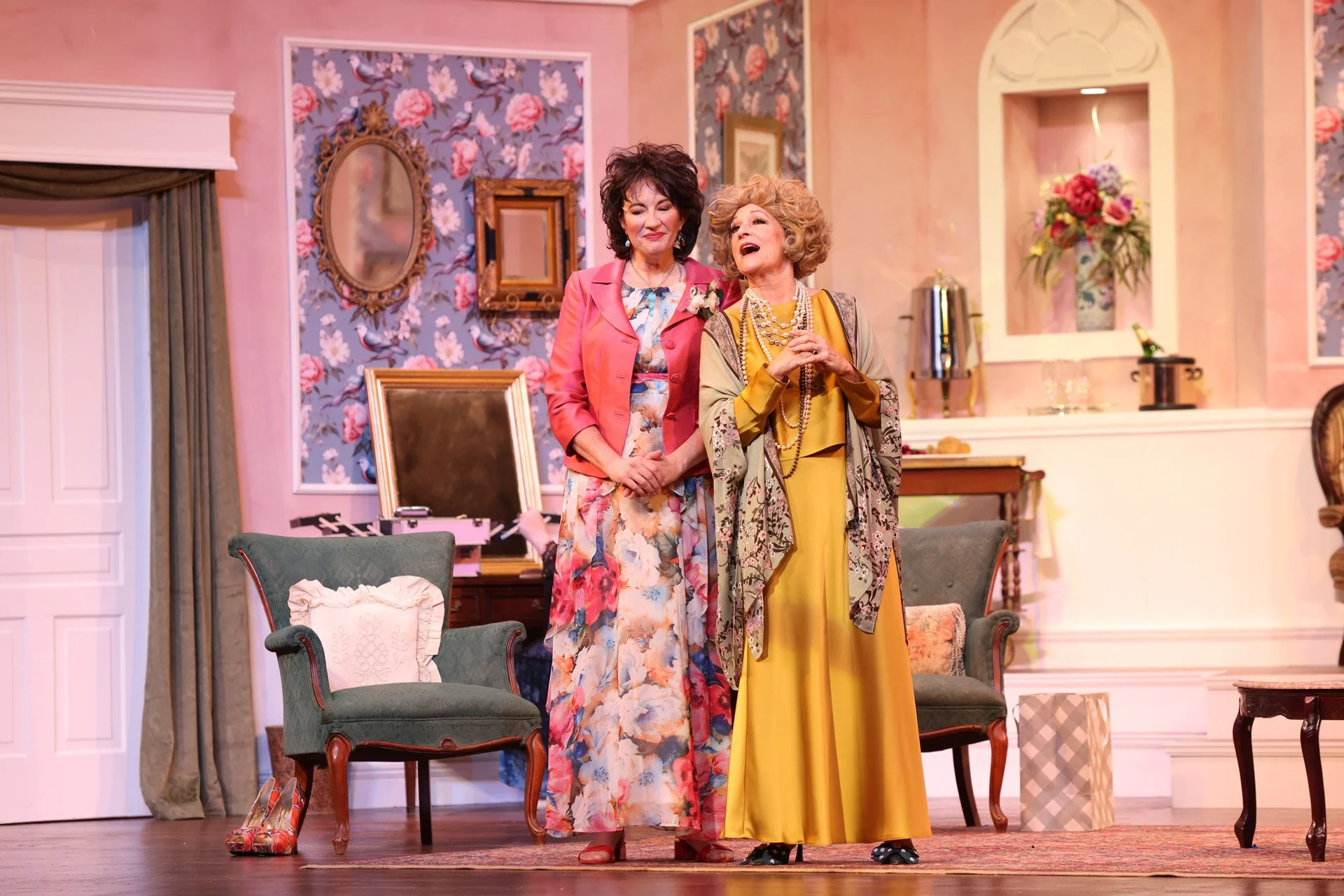 Two women on stage engaged in a theatrical performance in a room decorated with floral wallpaper, framed mirrors, and vintage furniture, one wearing a pink blazer and floral dress, the other in a yellow dress with layered necklaces. They are smiling and talking.