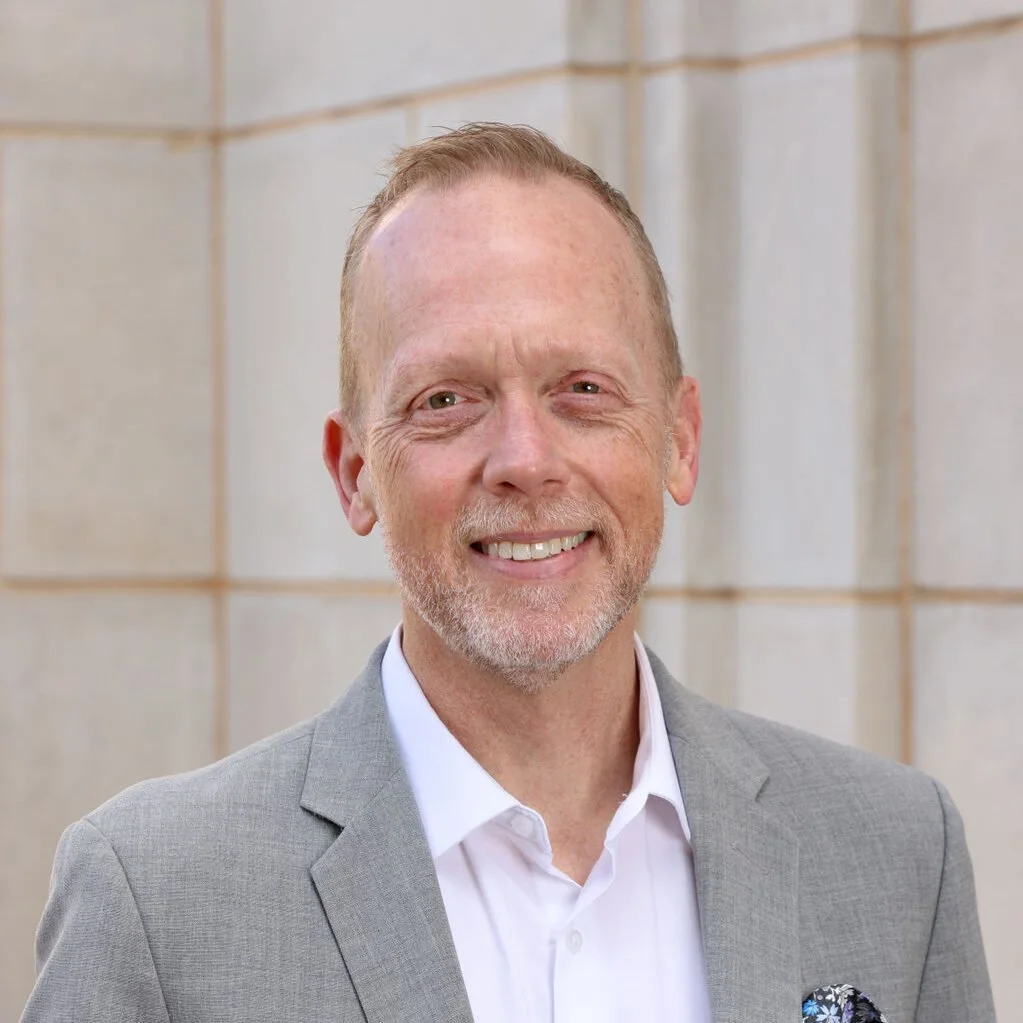 A middle-aged man with light skin, red hair, and a beard, smiling, wearing a light gray suit and white shirt, standing outdoors in front of a light-colored stone wall.