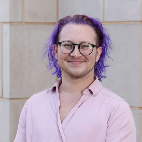 A young man with purple hair, glasses, and a light pink shirt, smiling in front of a beige tiled wall.