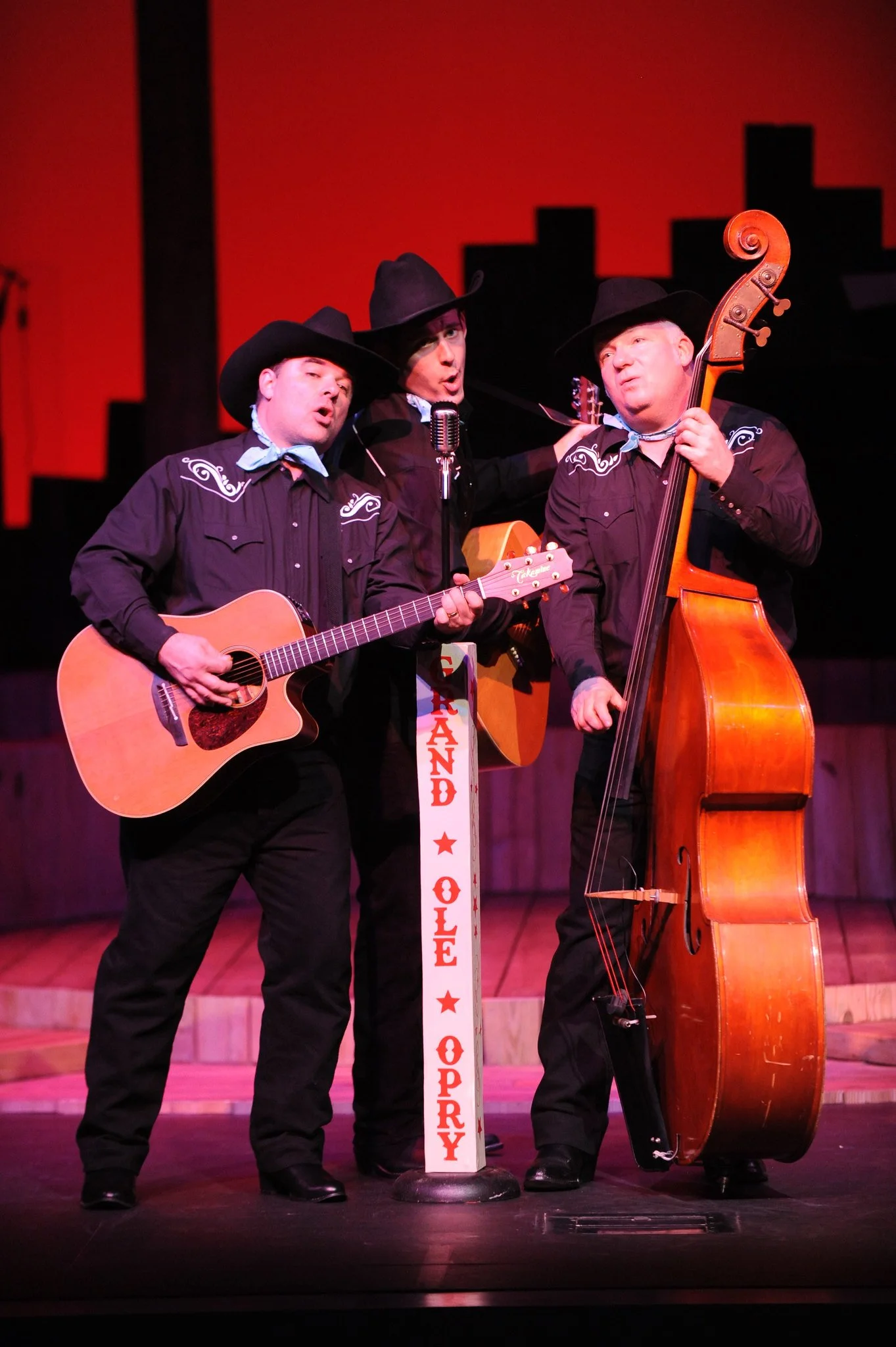 Three men dressed as cowboys singing and playing guitars on a stage with a city skyline silhouette in the background. The stage has a sign that reads 'Sunday Ole Opry'.