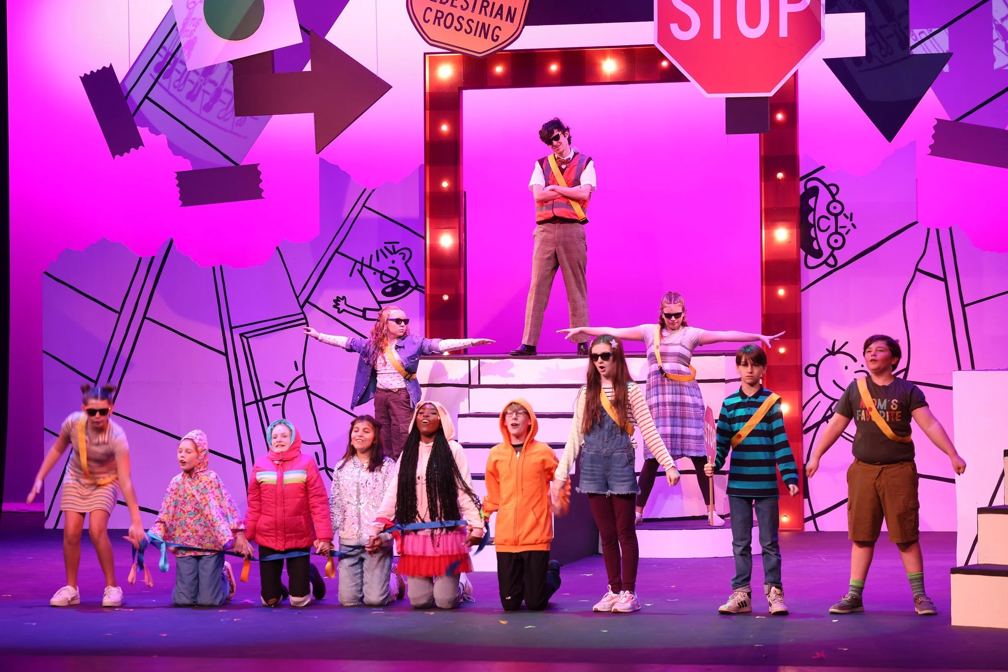 Group of children performing on stage with colorful background and signs including 'Pedestrian Crossing' and 'Stop' sign.