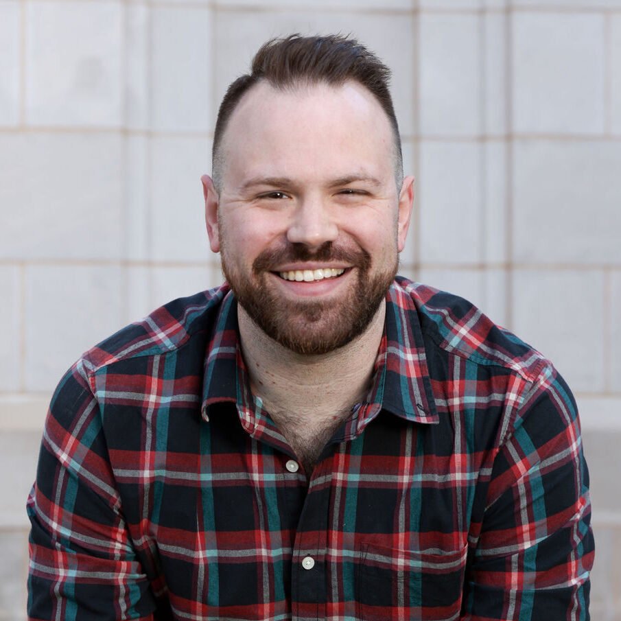 A smiling man with a beard and short hair, wearing a red, blue, and green plaid shirt, sitting against a light-colored tiled wall.