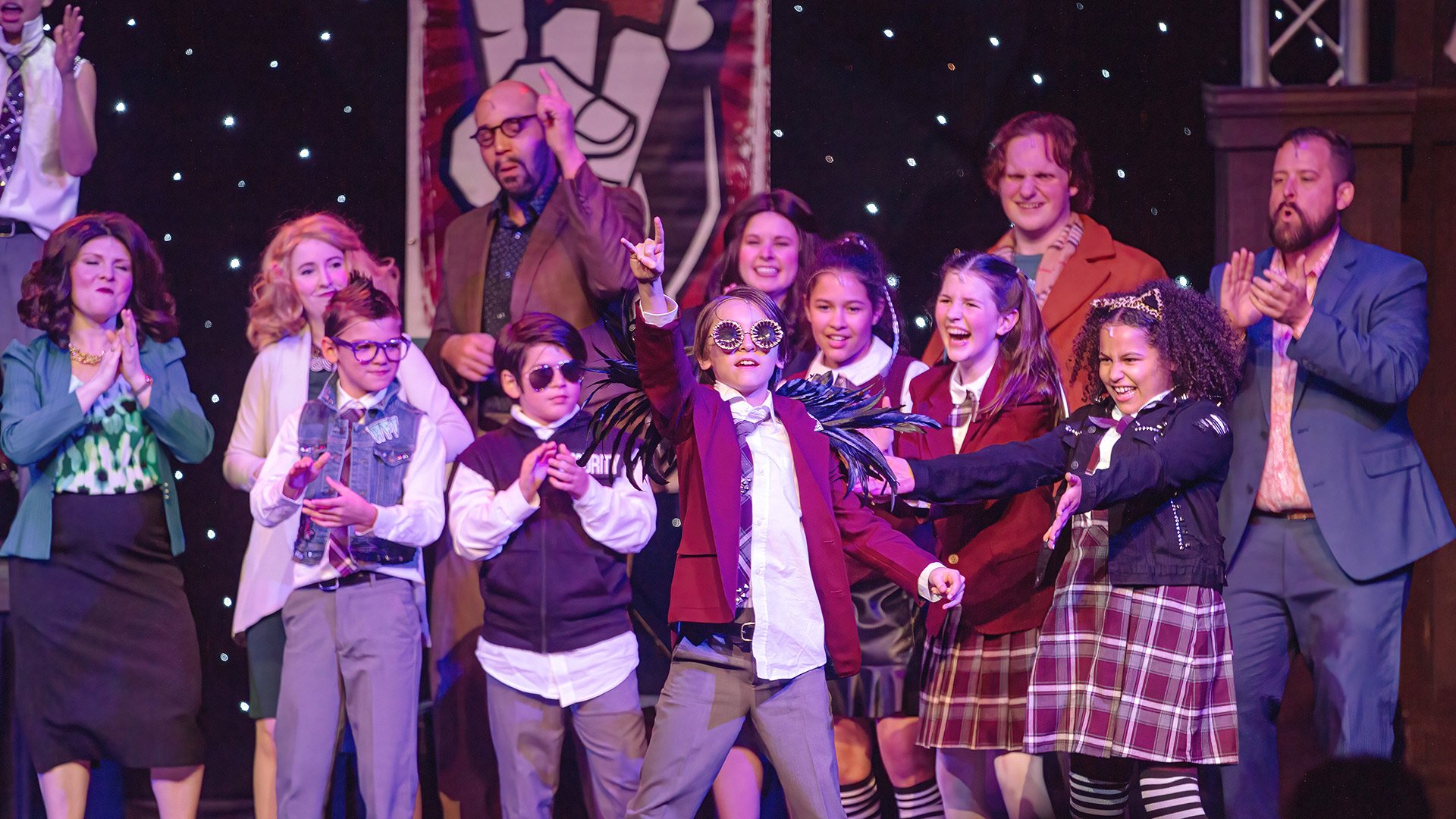 Group of children and adults dressed in school uniforms and costumes on stage, with a person in the middle wearing sunglasses and feathered accessories, participating in a theatrical performance or school play.