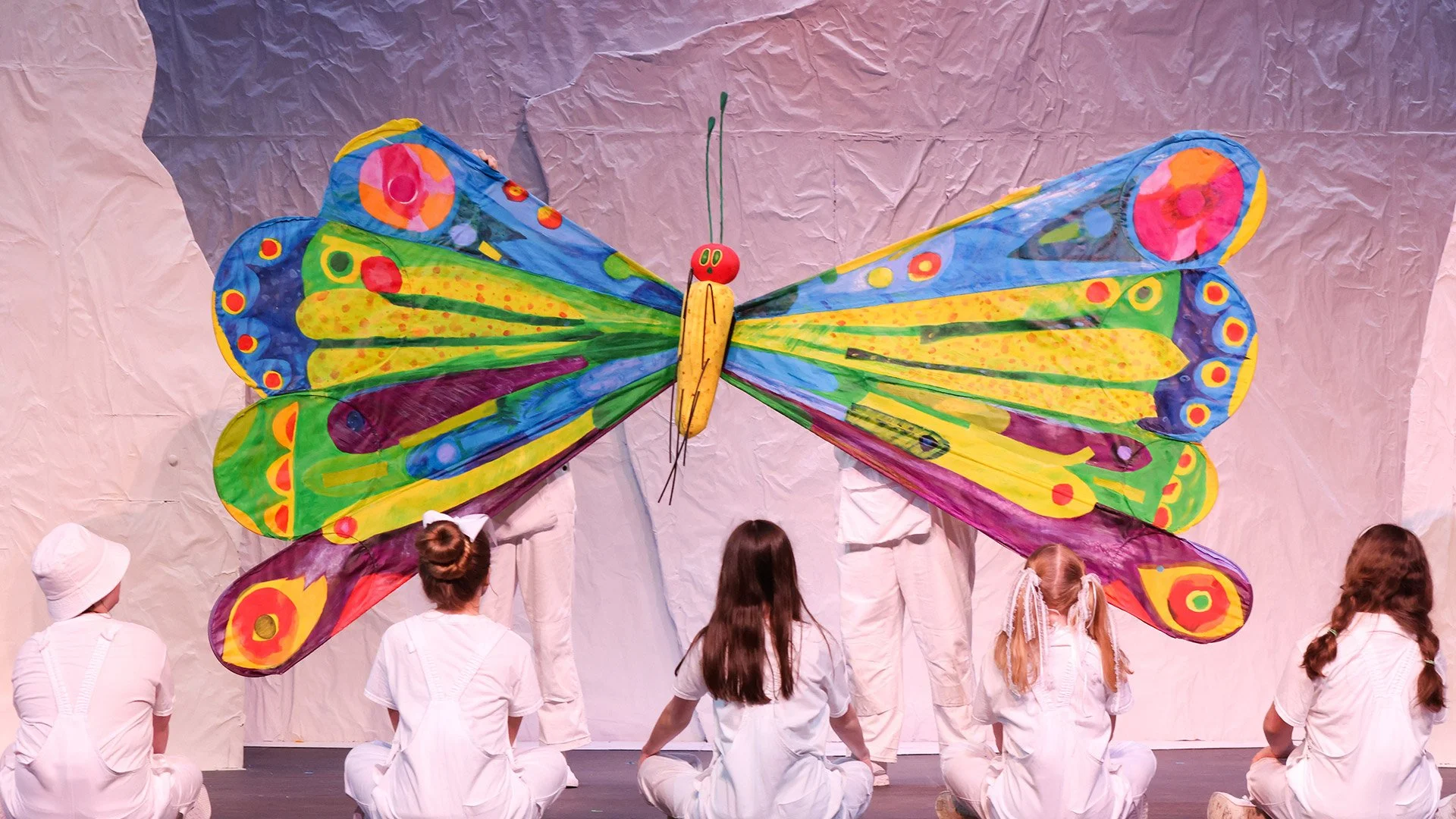 Children dressed in white kneeling and sitting in front of a large, colorful butterfly kite on stage at a performance or event.