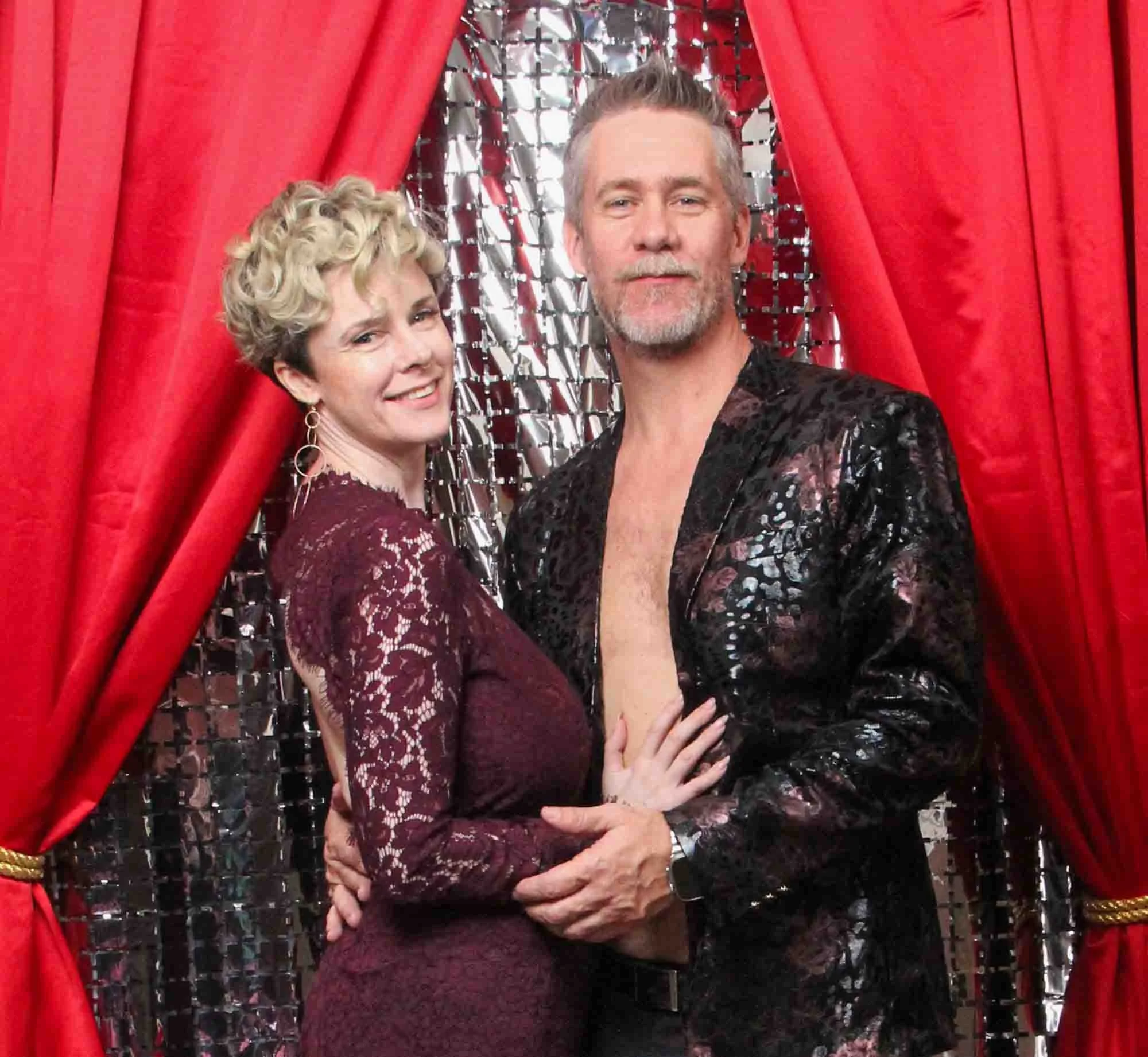 A smiling middle-aged woman and a middle-aged man pose together in front of a shiny sequined backdrop with red curtains on either side. The woman is wearing a burgundy lace dress with earrings, and the man is wearing an open, black patterned jacket.