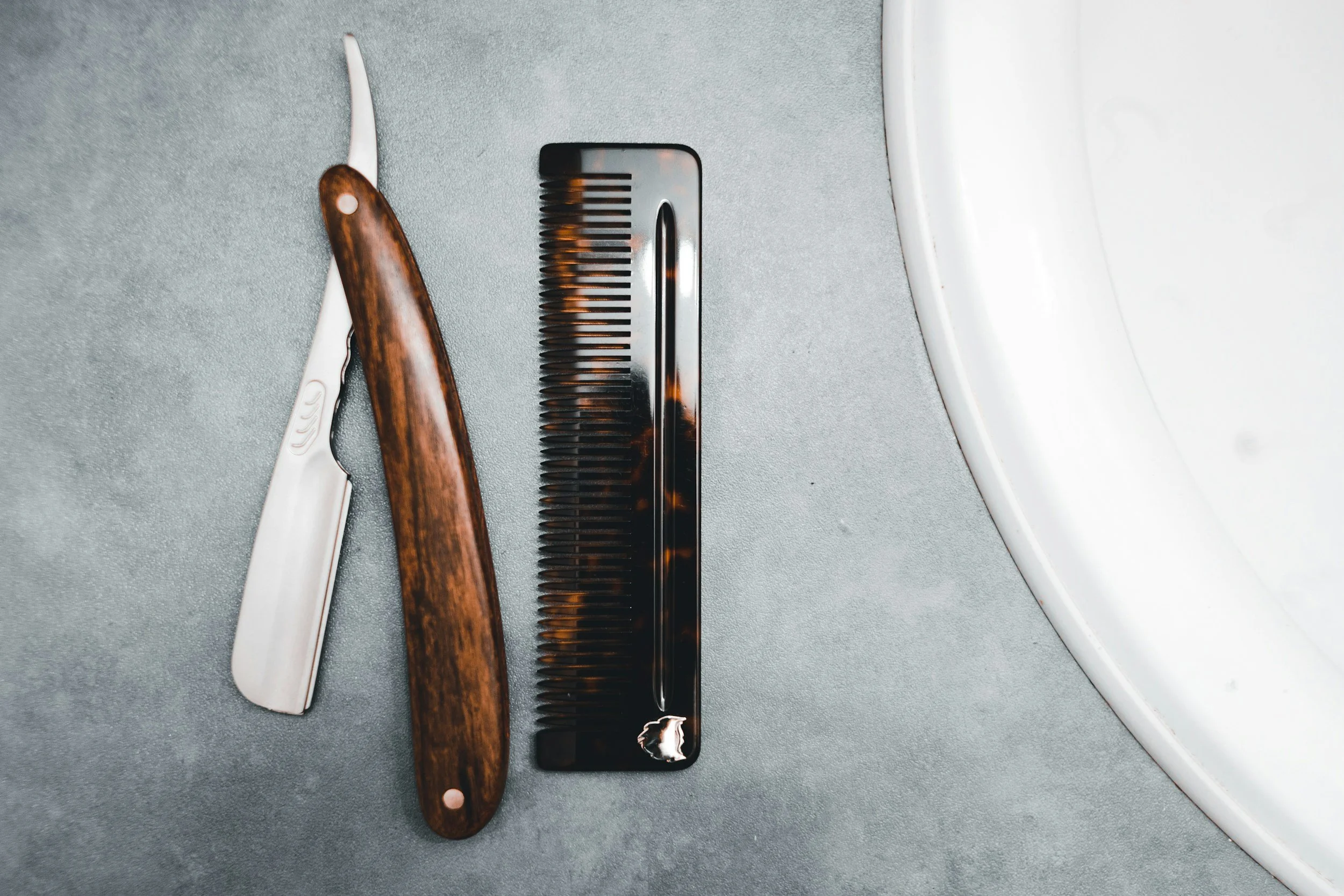 Safety razor, a wooden-handled straight razor, and a black comb on a gray surface near a white sink.