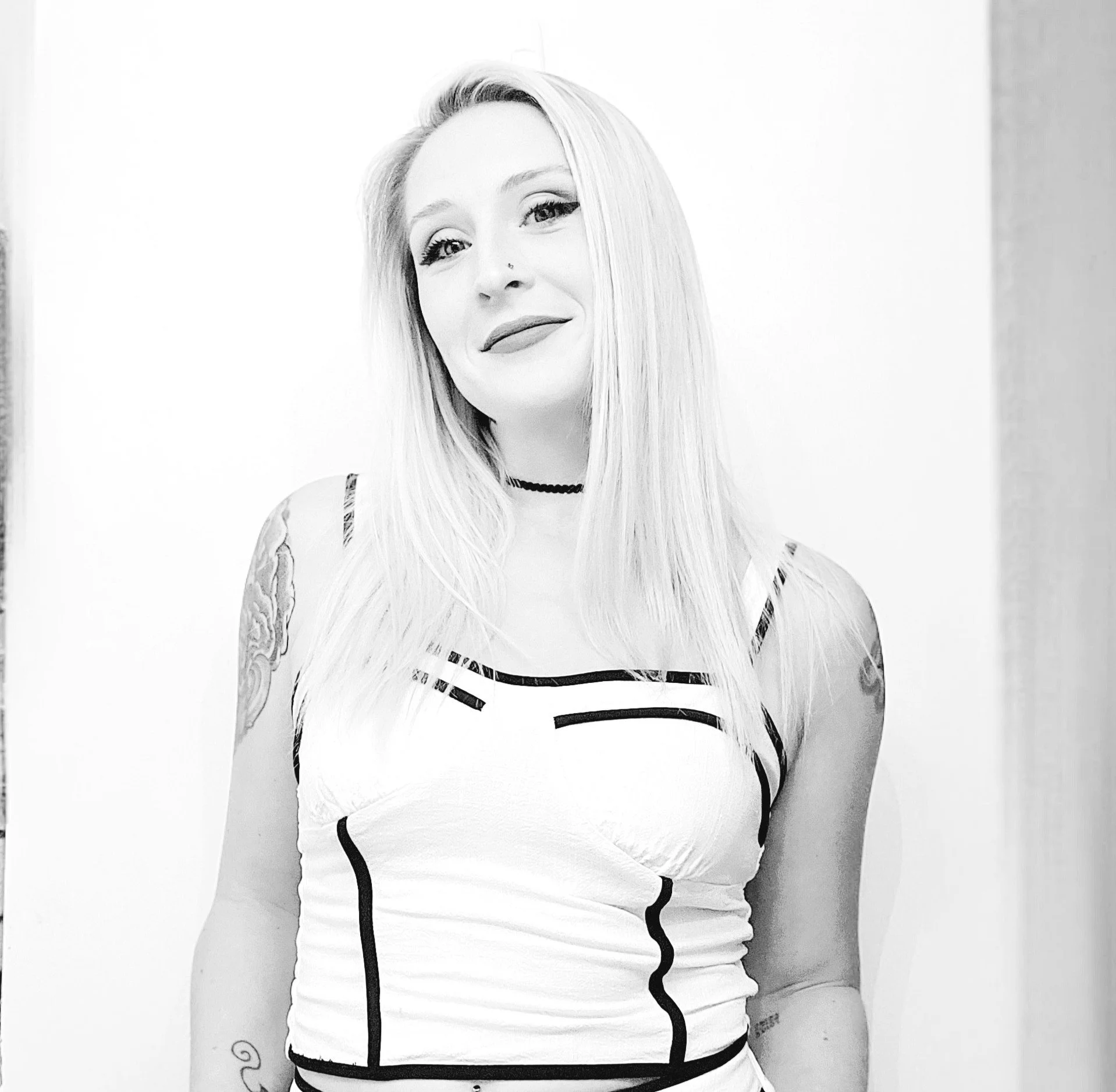 Black and white photo of a woman with long blonde hair, wearing a striped sleeveless top, a choker necklace, and tattoos on her arms, standing against a plain white background.