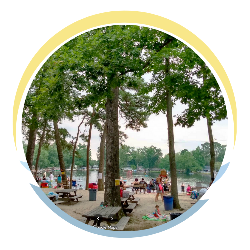 A lakeside park with trees, picnic tables, and people enjoying a day by the water.