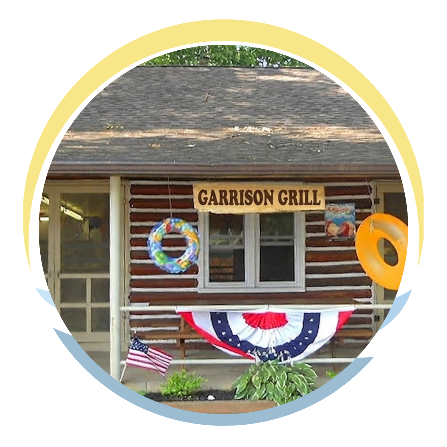 A small wooden building with a sign that says 'Snack Stand'. There are two inflatable rings, one blue with colorful decorations and the other orange, hanging in front of the building. An American flag is partially visible, and a patriotic bunting decoration is draped across the front. There are plants and flowers at the base of the building.