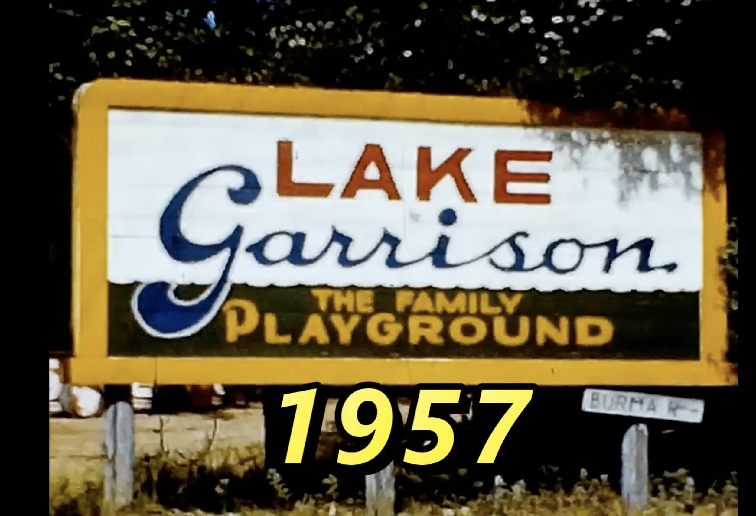 Sign for Lake Garrison, The Family Playground, dated 1957, with black, white, blue, red, and yellow colors, and a smaller street sign reading "Burbank" at the bottom right.