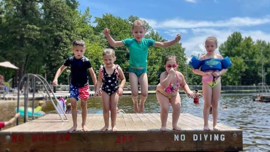 Five children in swimsuits playing and jumping off a wooden dock into a lake on a sunny day, with trees and a cloudy sky in the background.