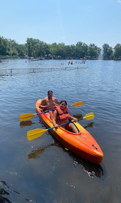 Two people kayaking on a lake, with trees and a clear sky in the background.