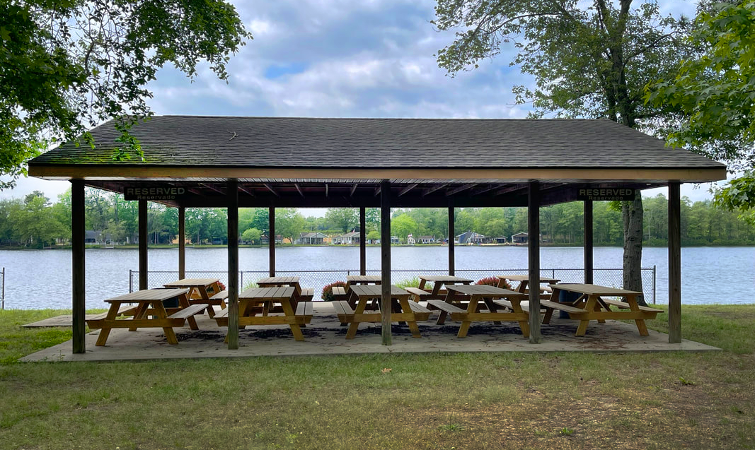 Covered picnic shelter with wooden tables by a lakeside, surrounded by trees and residential houses in the background.