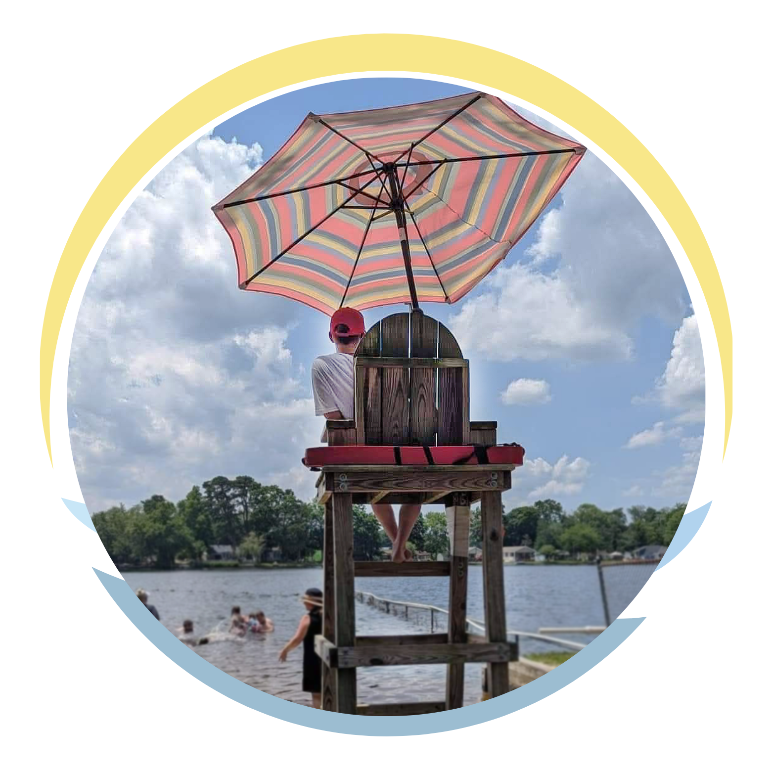 Person sitting on a lifeguard chair under a colorful striped umbrella by a lake, with people swimming and trees in the background on a sunny day.