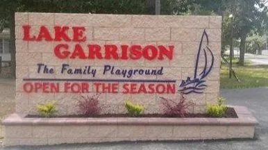 Sign for Lake Garrison, advertising as a family playground and open for the season, with a sailboat graphic and small plants at the base.