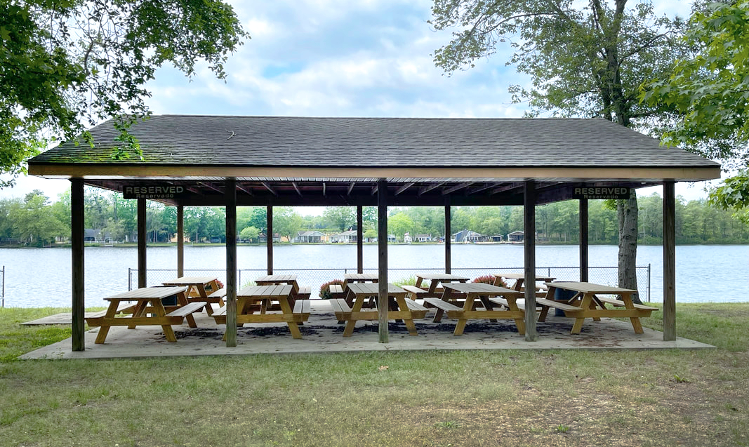 Wooden picnic shelter with tables near a lake, surrounded by trees, under a partly cloudy sky.