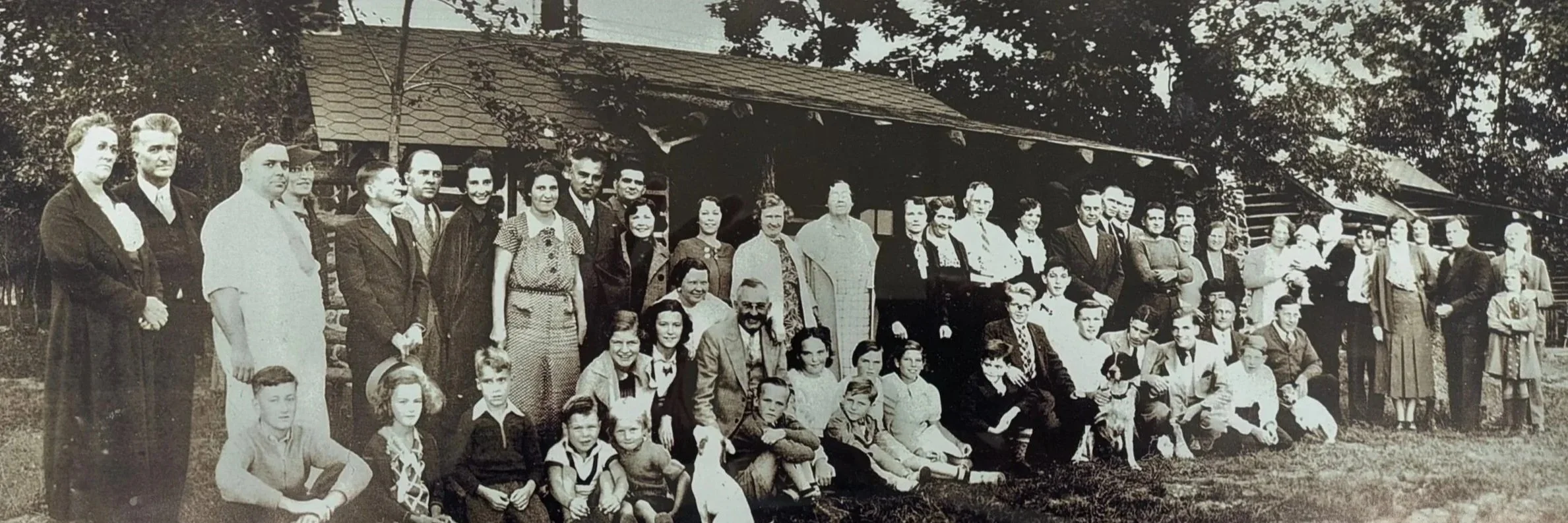 A large group of people, including children and adults, posed outdoors in front of a building with a shingled roof, during daytime. The photo is in black and white.