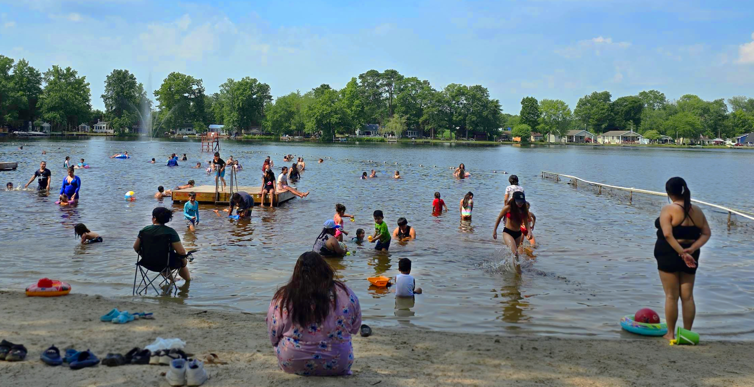People swimming and playing in a lake on a sunny day with trees and houses in the background.