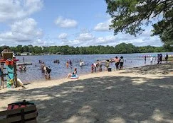 People enjoying a day at the beach by a lake with boats and trees in the background.
