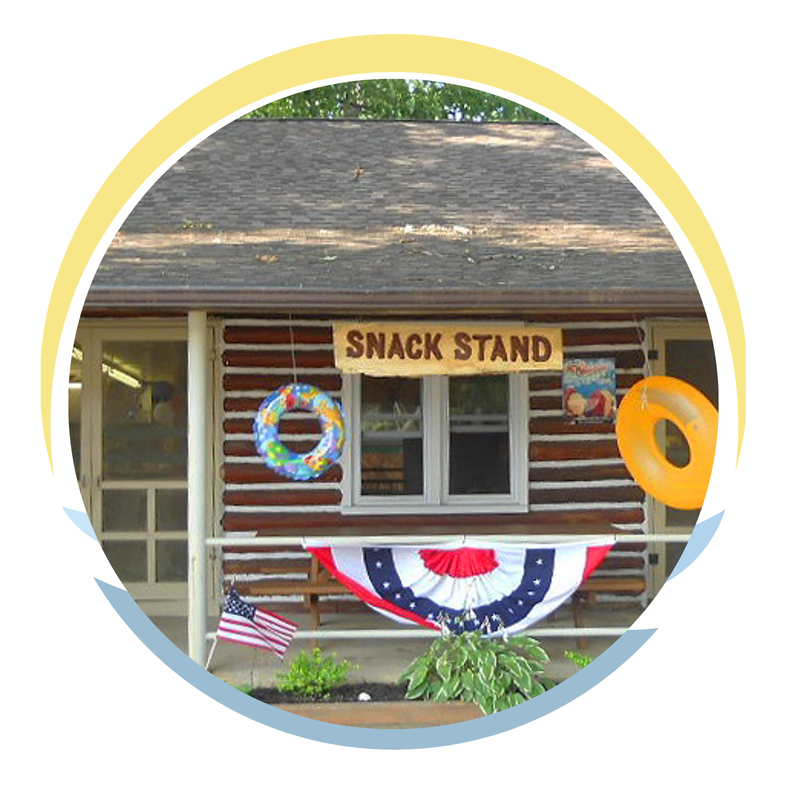 A small wooden building with a sign that says 'Snack Stand'. There are two inflatable rings, one blue with colorful decorations and the other orange, hanging in front of the building. An American flag is partially visible, and a patriotic bunting decoration is draped across the front. There are plants and flowers at the base of the building.