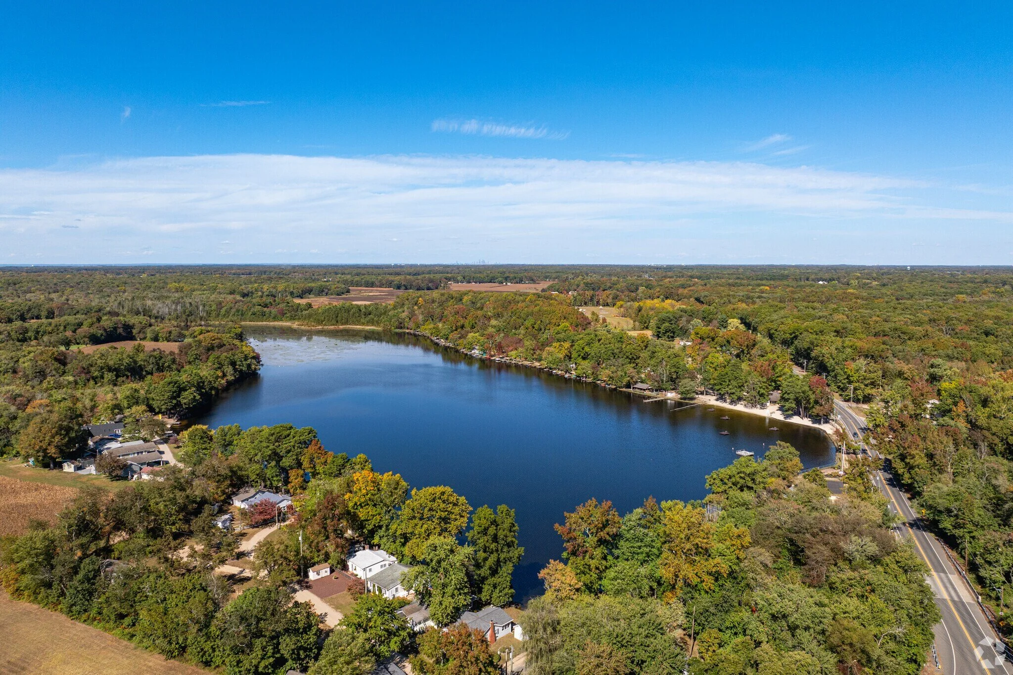 Aerial view of a small lake surrounded by trees with colorful autumn foliage, residential houses near the water, and a road on the right side, under a blue sky with scattered clouds.