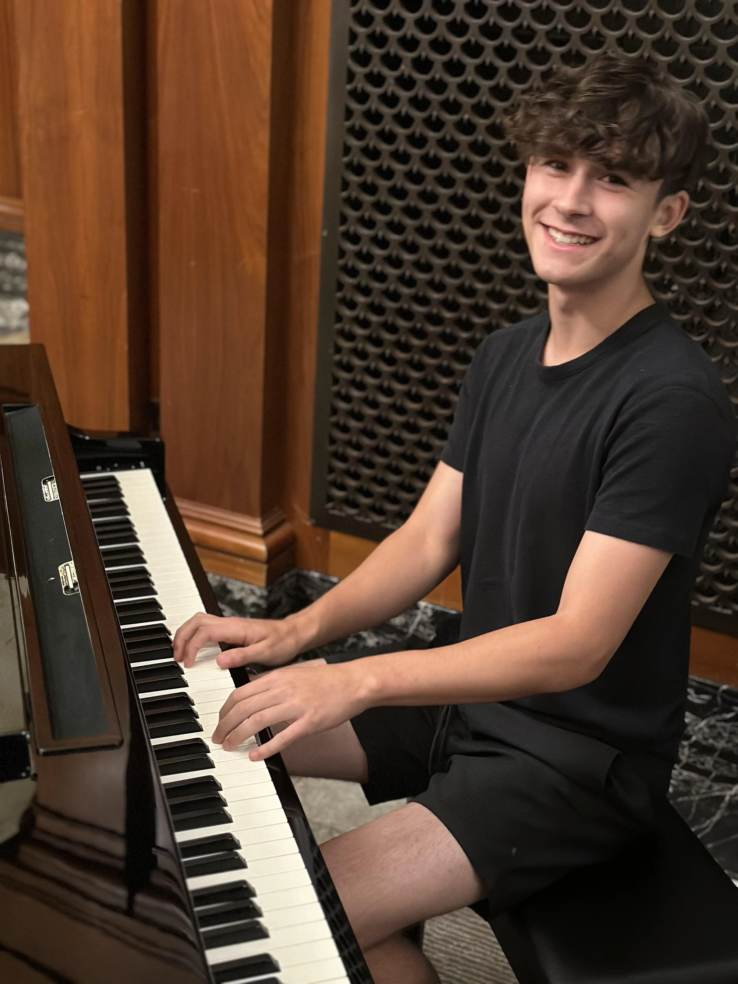 A young man with curly brown hair wearing a black t-shirt and black shorts, sitting at a piano, smiling at the camera in a music studio or practice room.