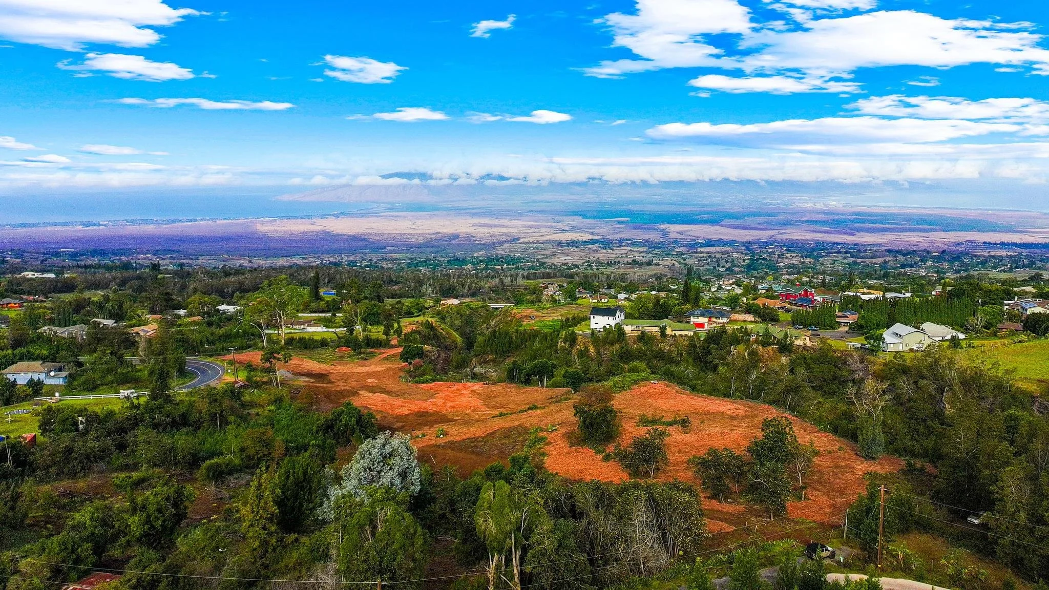 A scenic aerial view of a suburban area with houses, trees, and open land, with mountains and a body of water in the distance under a partly cloudy sky.