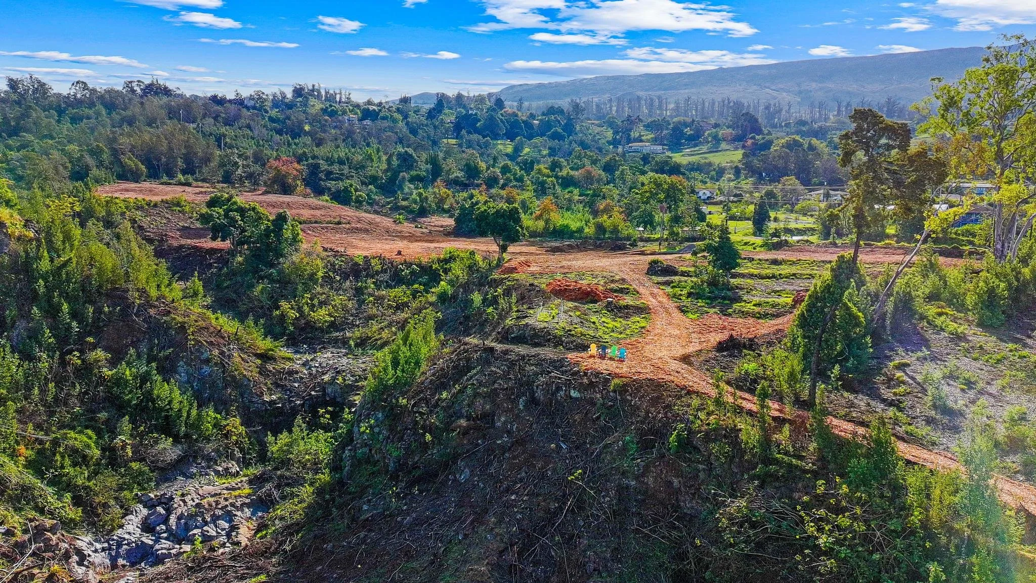 Landscape with trees, dirt paths, a small waterfall, and houses in the distance under a blue sky with clouds.