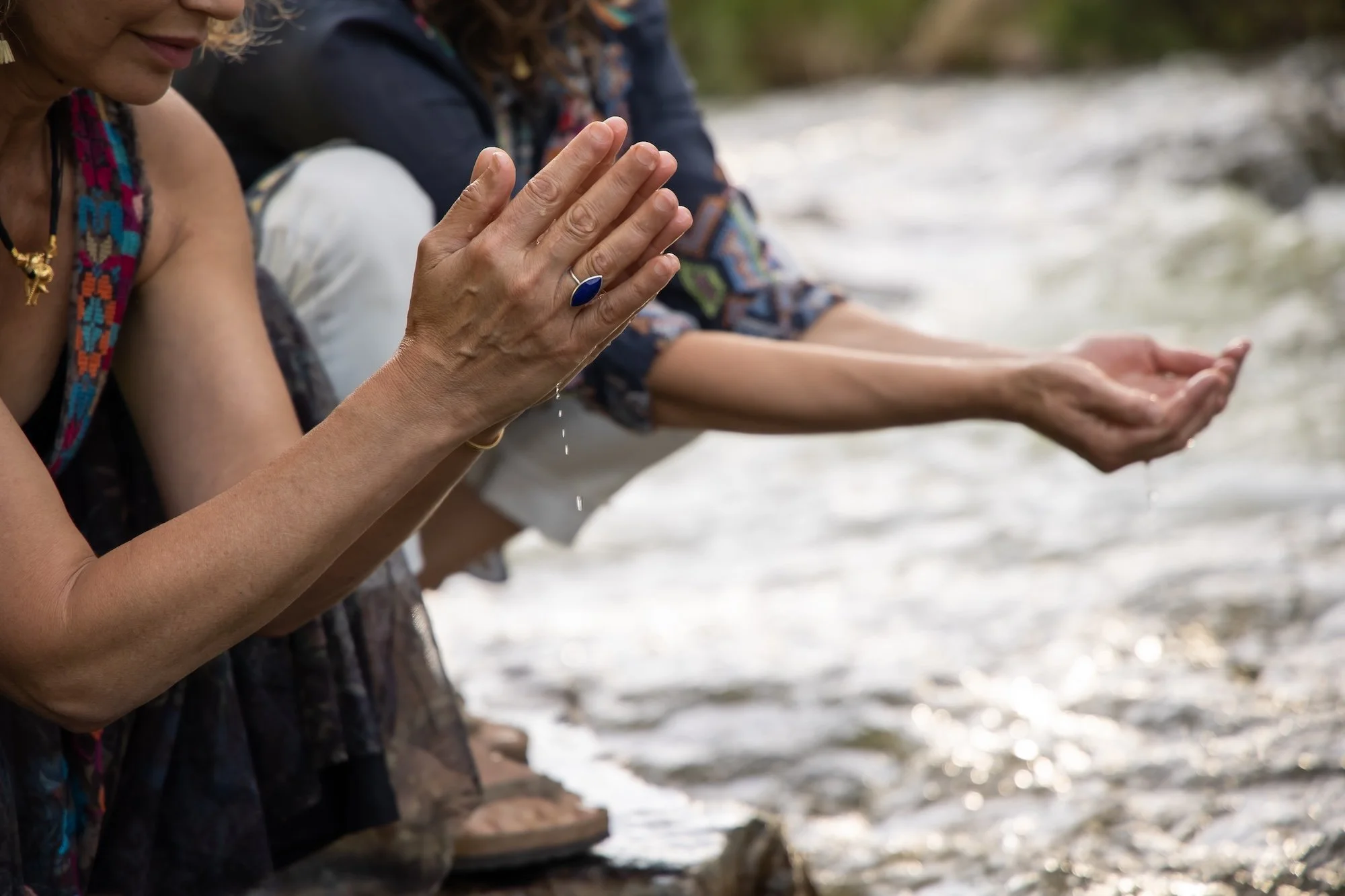 Two people on a riverbank with their hands extended over the water, performing a ritual. One woman is wearing a colorful beaded necklace and a large blue ring. Water is flowing over rocks in the background.