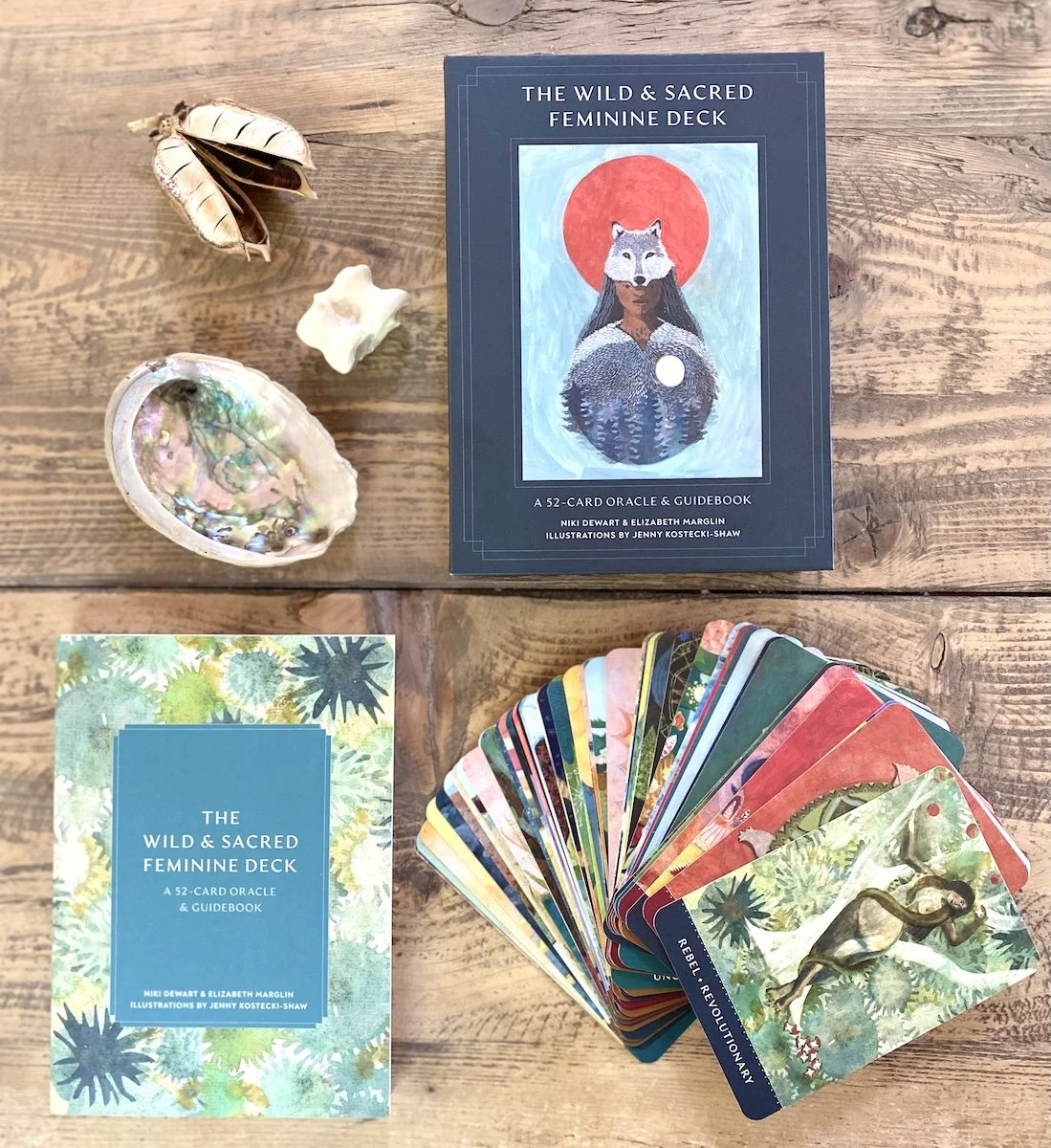 A deck of tarot or oracle cards titled 'The Wild & Sacred Feminine Deck' with a blue cover featuring an illustrated woman with a wolf face and a red sun behind her amongst found nature objects, all arranged on a wooden surface.