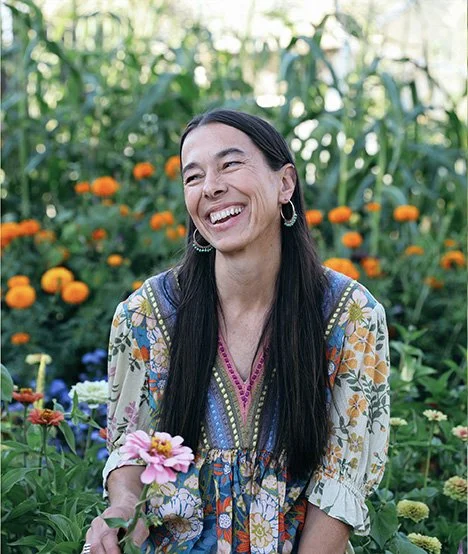 Woman with long dark hair smiling in a garden with colorful flowers and green foliage.