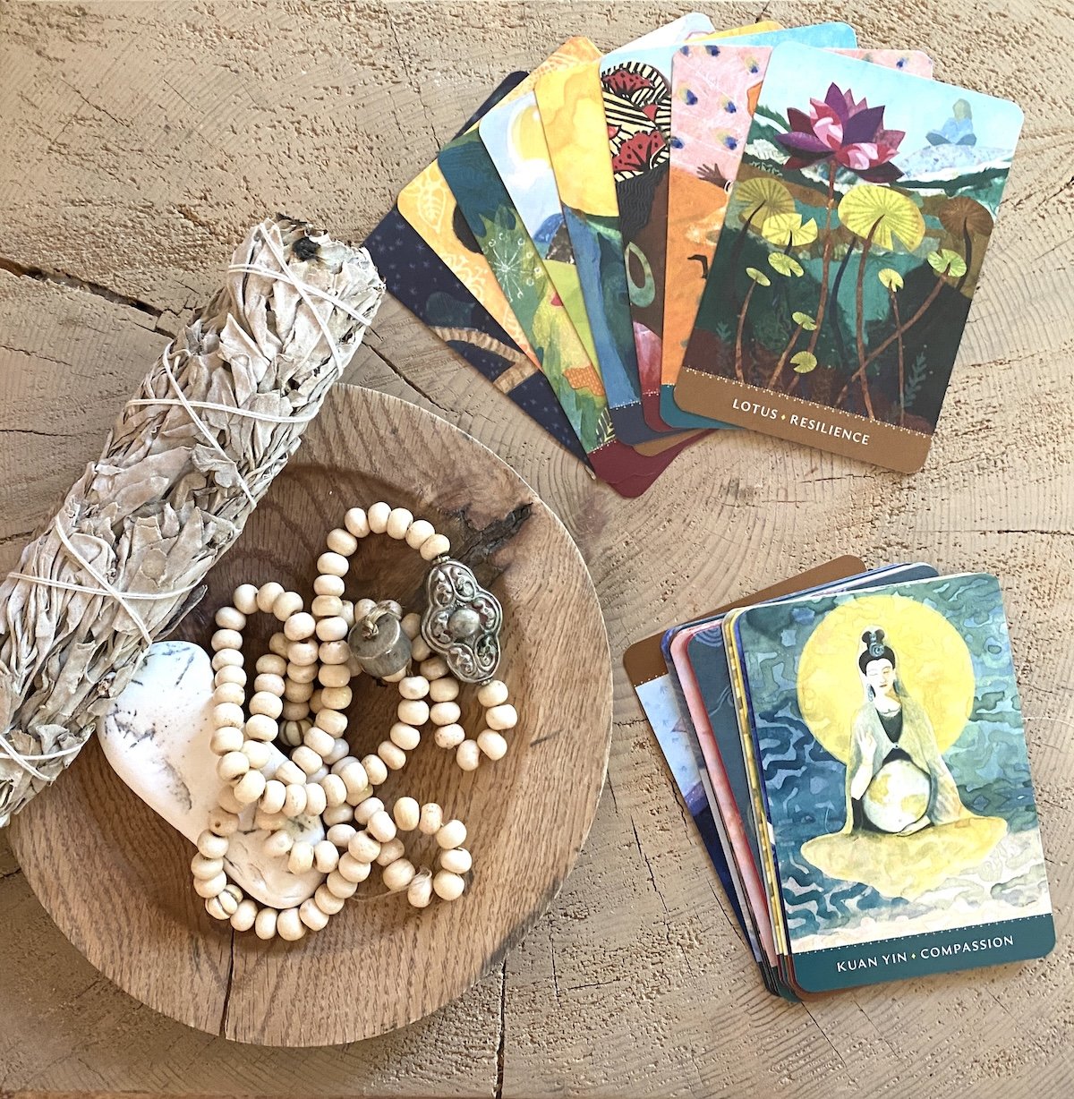 A round wooden tray with sage bundle, wooden bead necklaces, a silver charm, and a white stone two stacks of colorful tarot cards with artistic illustrations, including one titled 'Compassion' and another labeled 'Resilience' featuring lotus flowers.