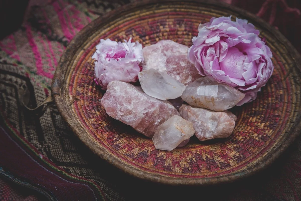 Pink peony flowers and rose quartz crystals in a woven basket on decorative fabric.