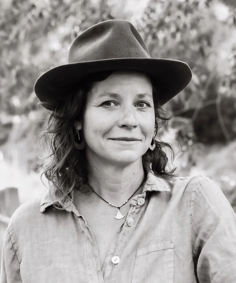 A woman with dark wavy hair wearing a wide-brimmed hat, earrings, a necklace, and a button-up shirt, outdoors with blurred trees in the background.
