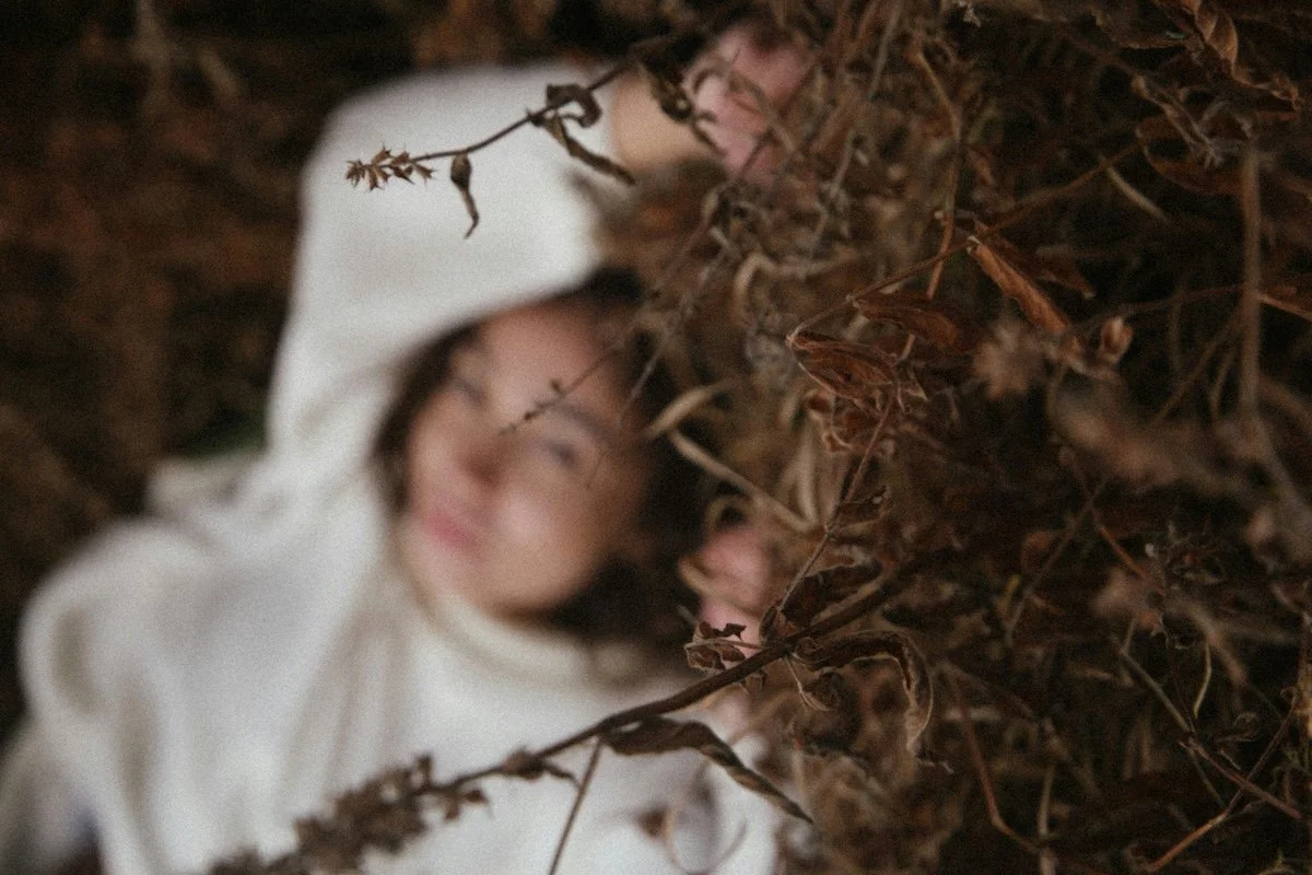 A woman lying among leaves and twigs, with her head slightly tilted and looking at the camera.
