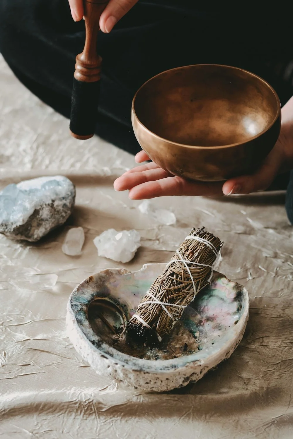 A person holding a brass singing bowl with their left hand and a wooden mallet with a black handle in their right hand, placed on a textured surface with crystals, a smudge stick, and a shell-shaped bowl.