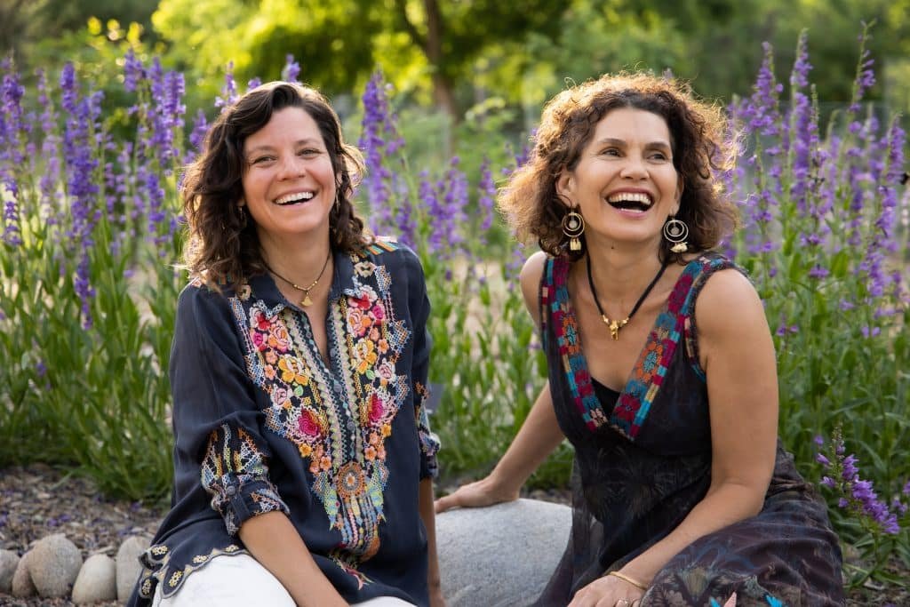 Two women sitting outdoors among purple flowers, smiling and laughing in a garden setting.