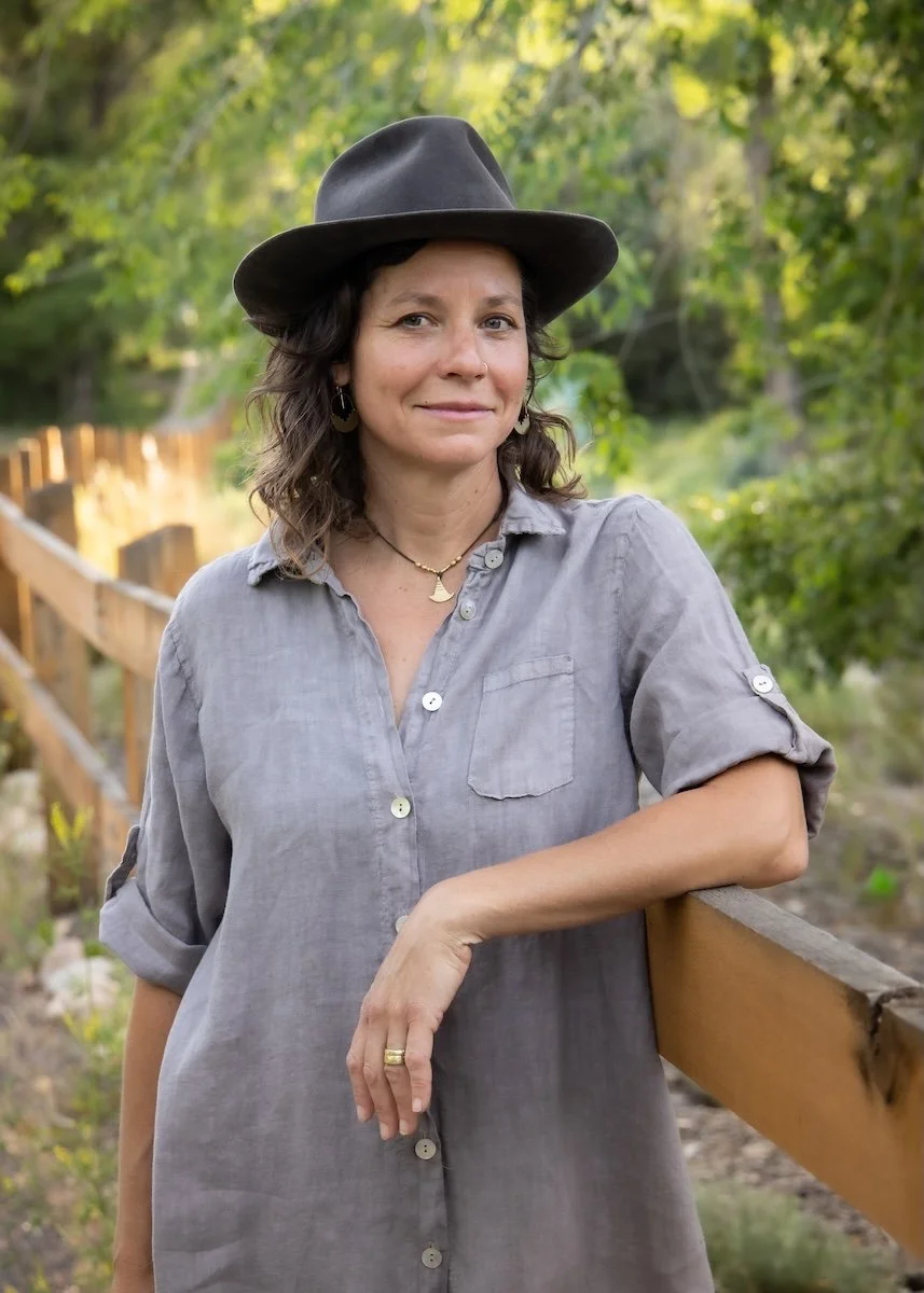 Woman with wavy dark brown hair wearing a gray button-up shirt and a wide-brim black hat, standing outdoors by a wooden fence surrounded by trees and greenery.