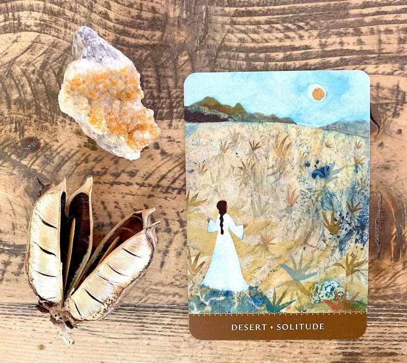 An illustrated card featuring desert landscape with mountains and a woman in a white dress, alongside a gemstone, a butterfly, and a feather on a wooden surface.