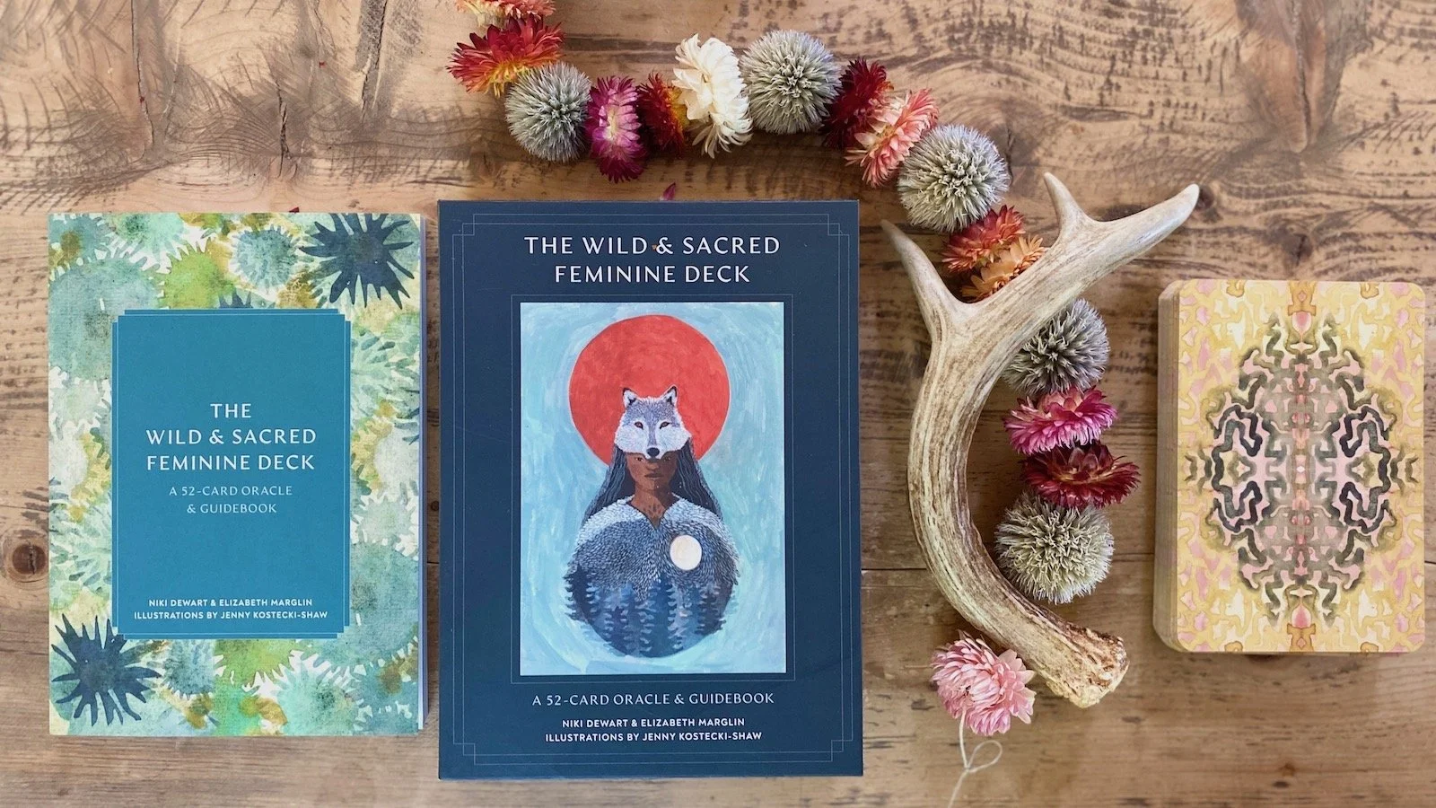 A collection of an oracle deck & guidebook titled 'The Wild & Sacred Feminine Deck' placed on a wooden table, accompanied by a floral crown, a deer antler, and cards with an intricate patterned design.