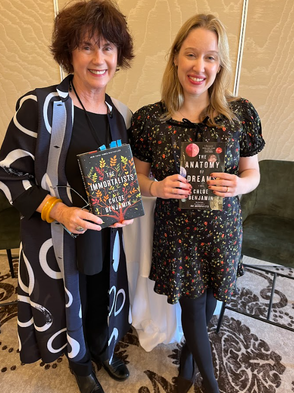 Two women smiling and holding books, standing indoors against a beige wall. The woman on the left holds a copy of the book "The Immortalists" by Chloe Benjamin, while the woman on the right holds "The Anatomy of Dreams" by Chloe Benjamin.