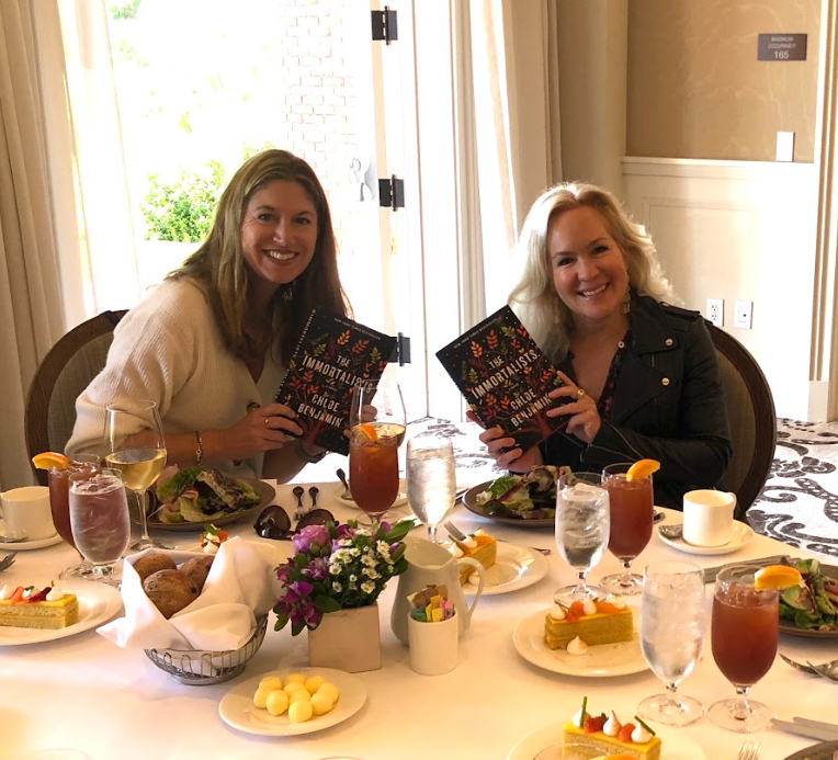 Two women sitting at a table with food and drinks, holding copies of a book titled 'The Immortalists.'