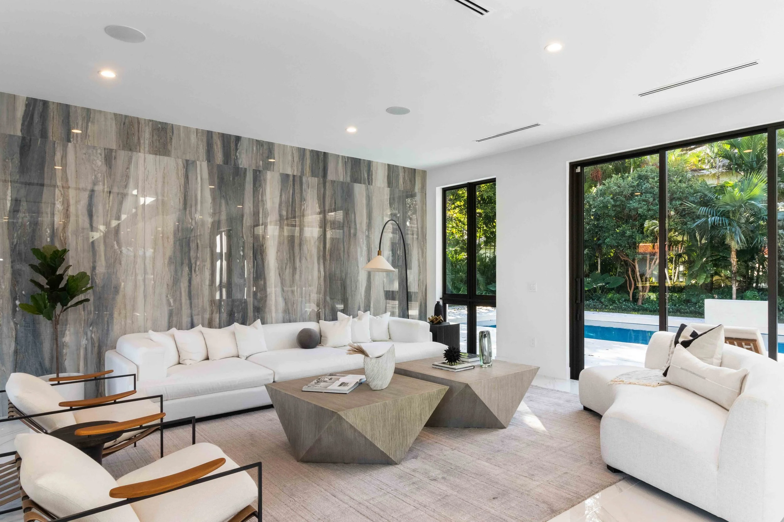 Contemporary living room with white sofas, faceted wood coffee tables, black metal lounge chairs, and dramatic gray stone accent wall overlooking the pool in Coral Gables, Interior Design by CURATED.