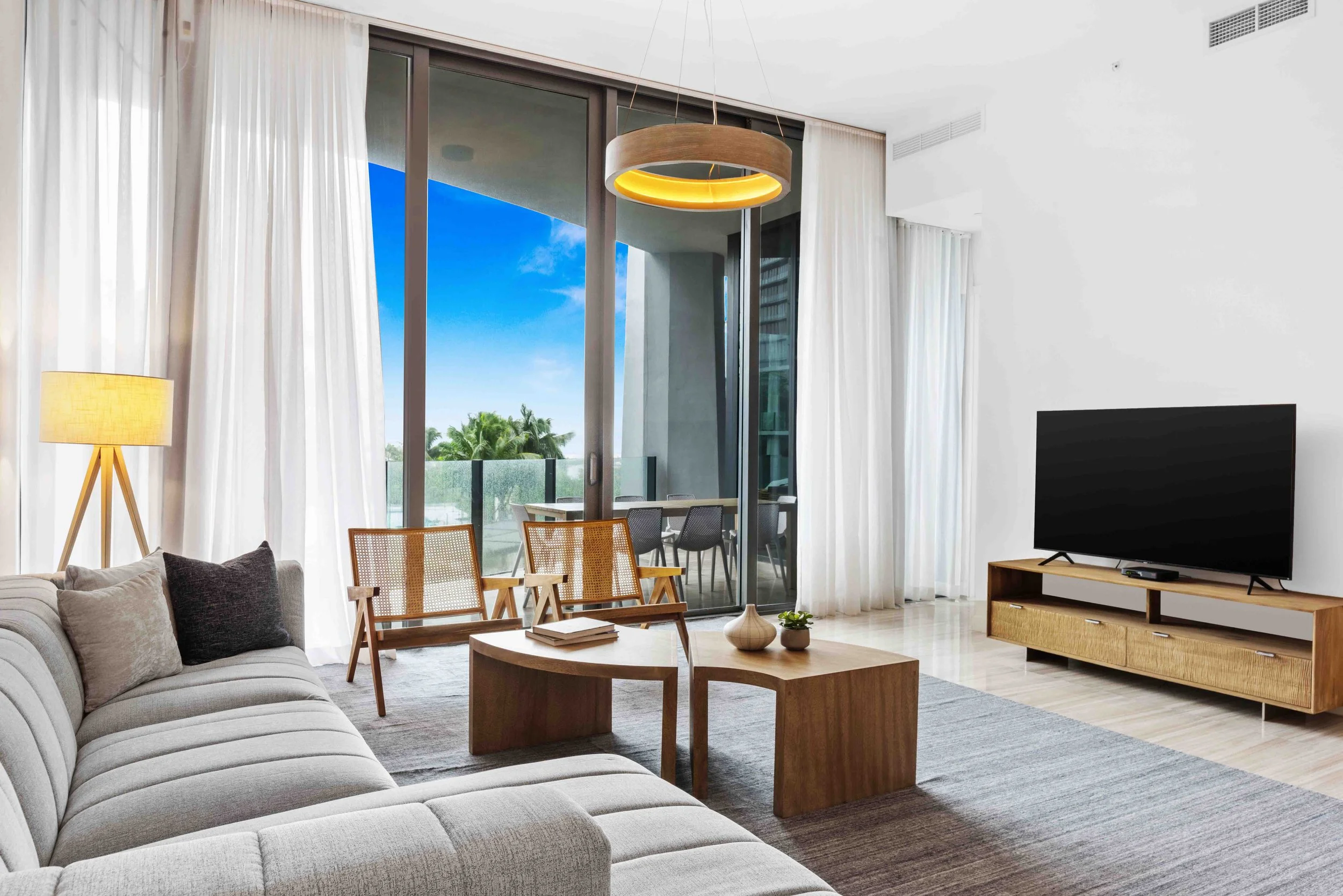 Mid-century coastal living room with channel-tufted gray sofa, nesting wood coffee tables, cane accent chairs, and tripod floor lamp in Coconut Grove, Interior Design by CURATED.