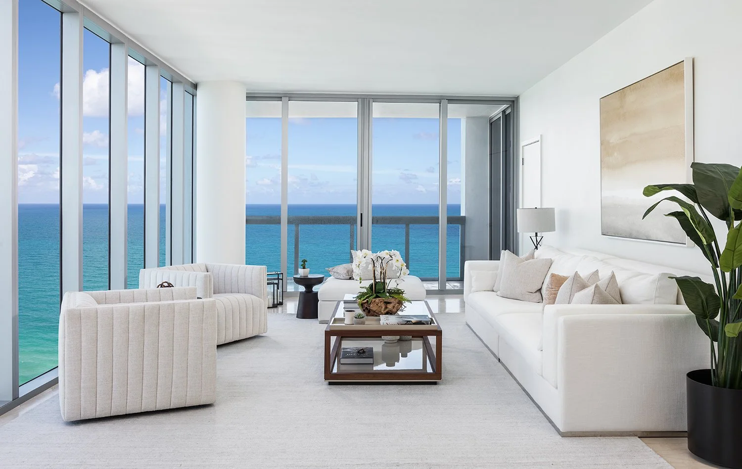 Oceanfront living room with white sofa, channel-tufted armchairs, wood and glass coffee table, and wraparound floor-to-ceiling ocean views, Interior Design by CURATED.