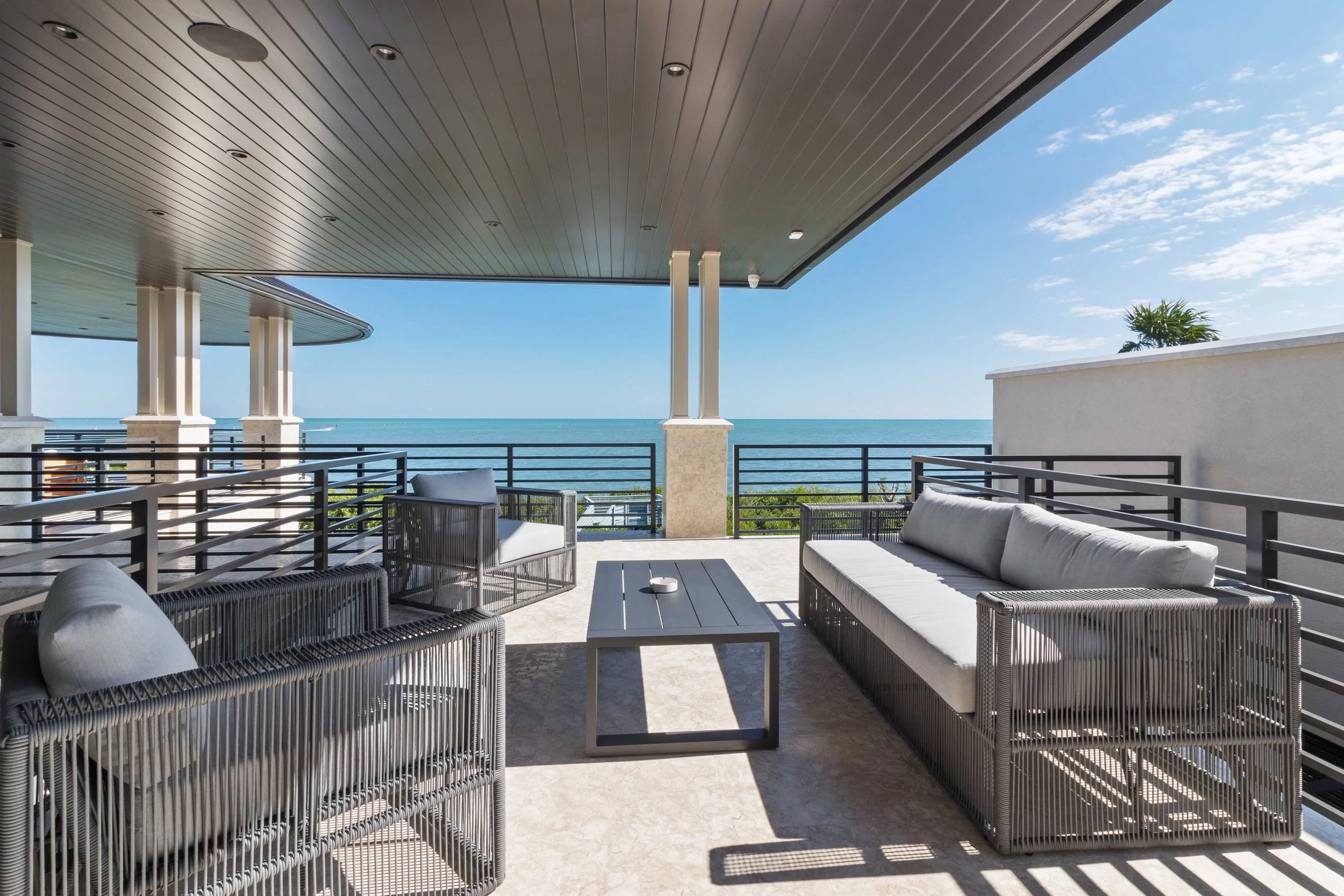 Covered oceanfront terrace with gray woven outdoor sofa and lounge chairs, slatted wood ceiling, and panoramic ocean views at Ocean Reef in Key Largo, Interior Design by CURATED.